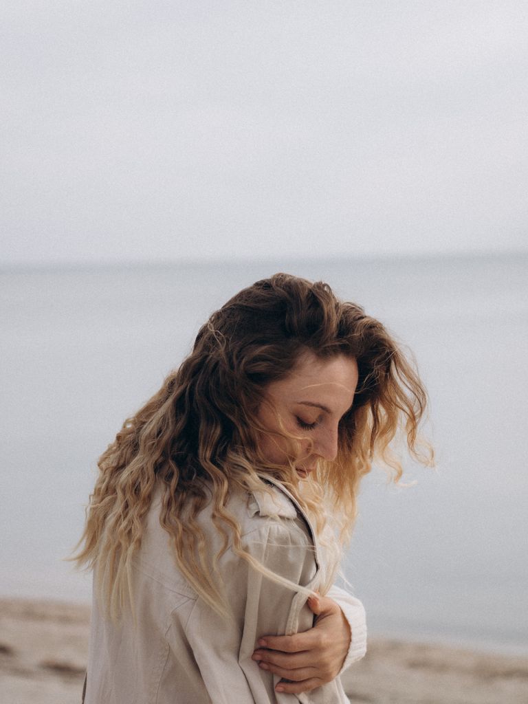 A woman in a beige jacket gazes pensively at a tranquil beach setting.