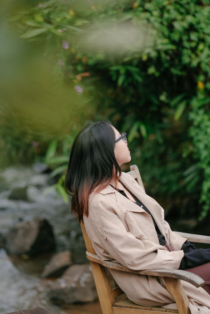 Woman in a beige coat sitting by a serene stream, surrounded by lush greenery.