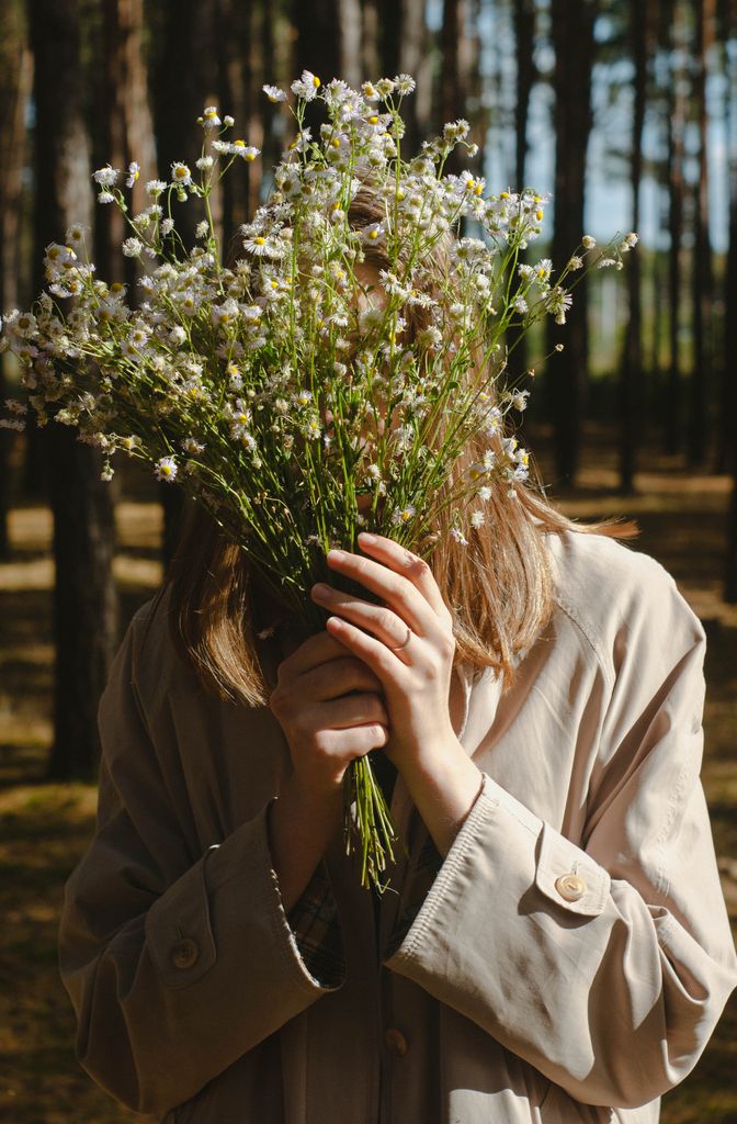 A woman hides her face with a bouquet of wildflowers in a sunlit forest.